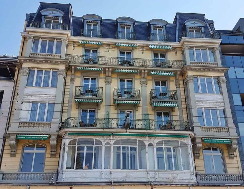Bright and elegant ironwork of a Haussmann-style hotel with wrought iron balconies and large traditional windows. Classic architecture, beige facade, lively street in Geneva.